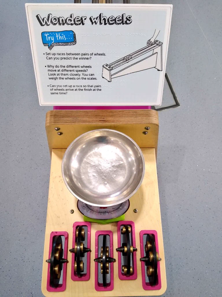 A light blue exhibit sign is shown on the top middle of the image, the title reads 'Wonder wheels'. Below this is a wooden table, with a silver tray for a set for weighing scales, and five black weighted wheels resting in pink rectangular slots.