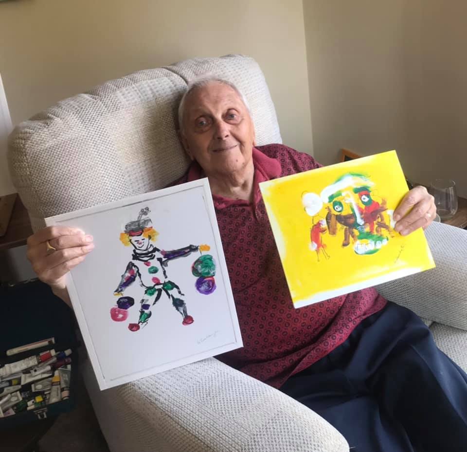 Harry sitting down holding up his clown and Sevilla paintings.