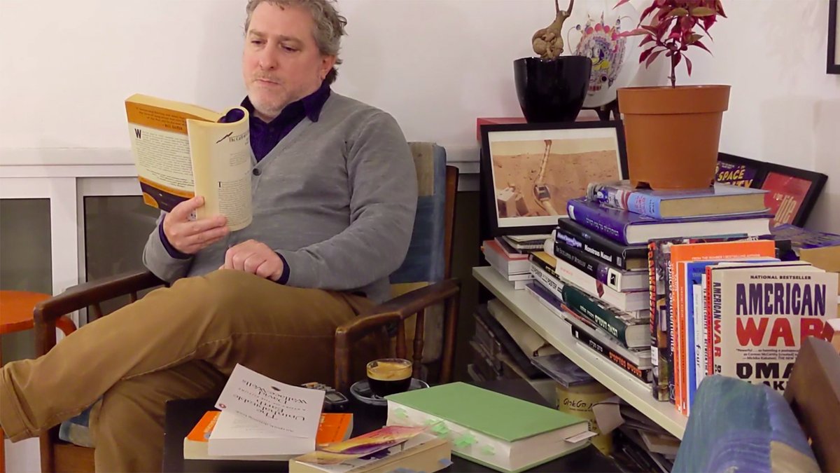 Uri Aviv sitzt ein Buch lesend in einem Sessel vor einem unübersichtlichen Bücherboard voller Bücher und vor einem kleinen Tisch auf dem ebenfalls Bücher und eine Kaffeetasse zu sehen sind. Screenshot aus der Session „Year(s) of the Uncanny“