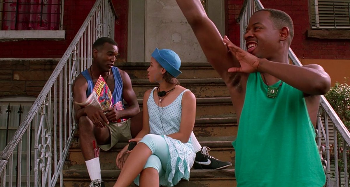 Three Black teens sit on the stoop of a building, hanging out. The boy in front is raising his arm to mime throwing a basketball, and his arm forms an arc that neatly frames the two kids behind him