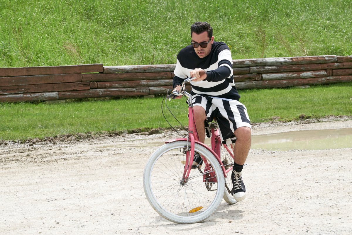 A series of three photos from the episode of Schitt's Creek where Mutt gifted Alexis a bright pink bike. In the first, Alexis (Annie Murphy) struggles to ride the bike as a new learner. In the second, David (Dan Levy) is also struggling with the bike. In the third, David is still on the bike and Alexis is trying to push him from behind. 