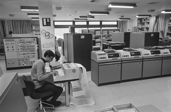A computer room from the Olympics. A person is typing on a large typewriter-like console. A console is behind with lights and buttons. At the right, four dishwasher-sized disk drives. Large computer boxes at the back.