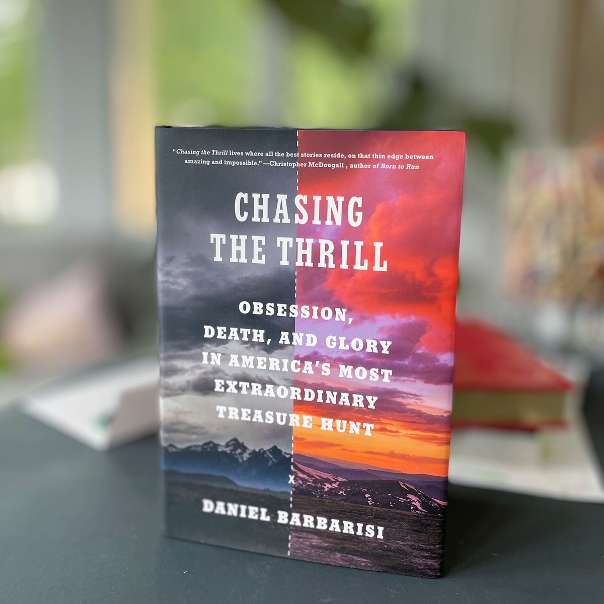 A copy of Chasing the Thrill by Daniel Barbarisi is standing faced out on a table with blurred out books and other items in the background.