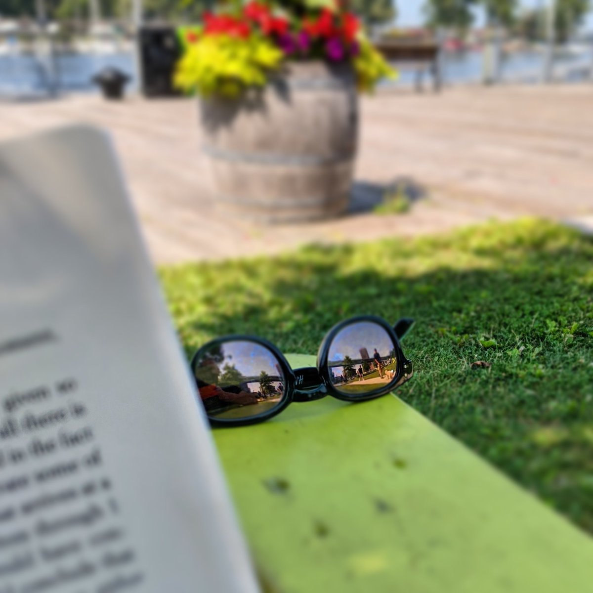 Looking over the pages of a book at a pair of mirror sunglasses balanced on the arm of an Adirondack chair showing the reflection of a person walking up behind me. In the distance is a boardwalk and river.