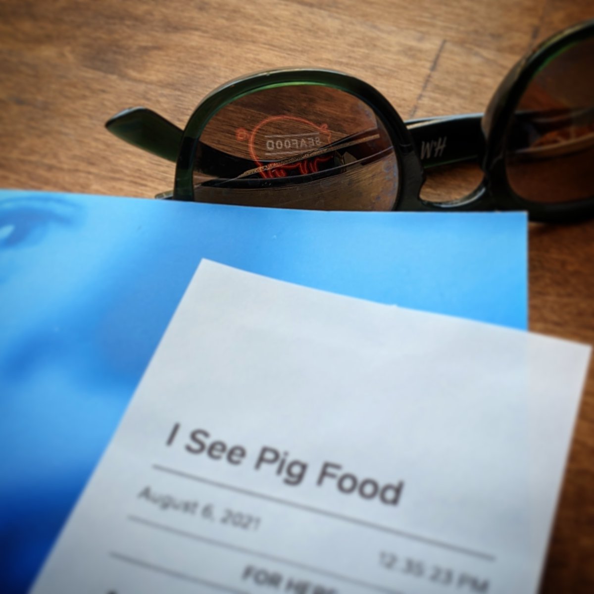 In the foreground is an order ticket with the customer name, I SEE PIG FOOD. Visible just behind it is a pair of reflective sunglasses, and in the reflection is a t-shirt with an image of a pink neon sign representing a profile of a pink pig with neon blue SEAFOOD printed across the length of its body.