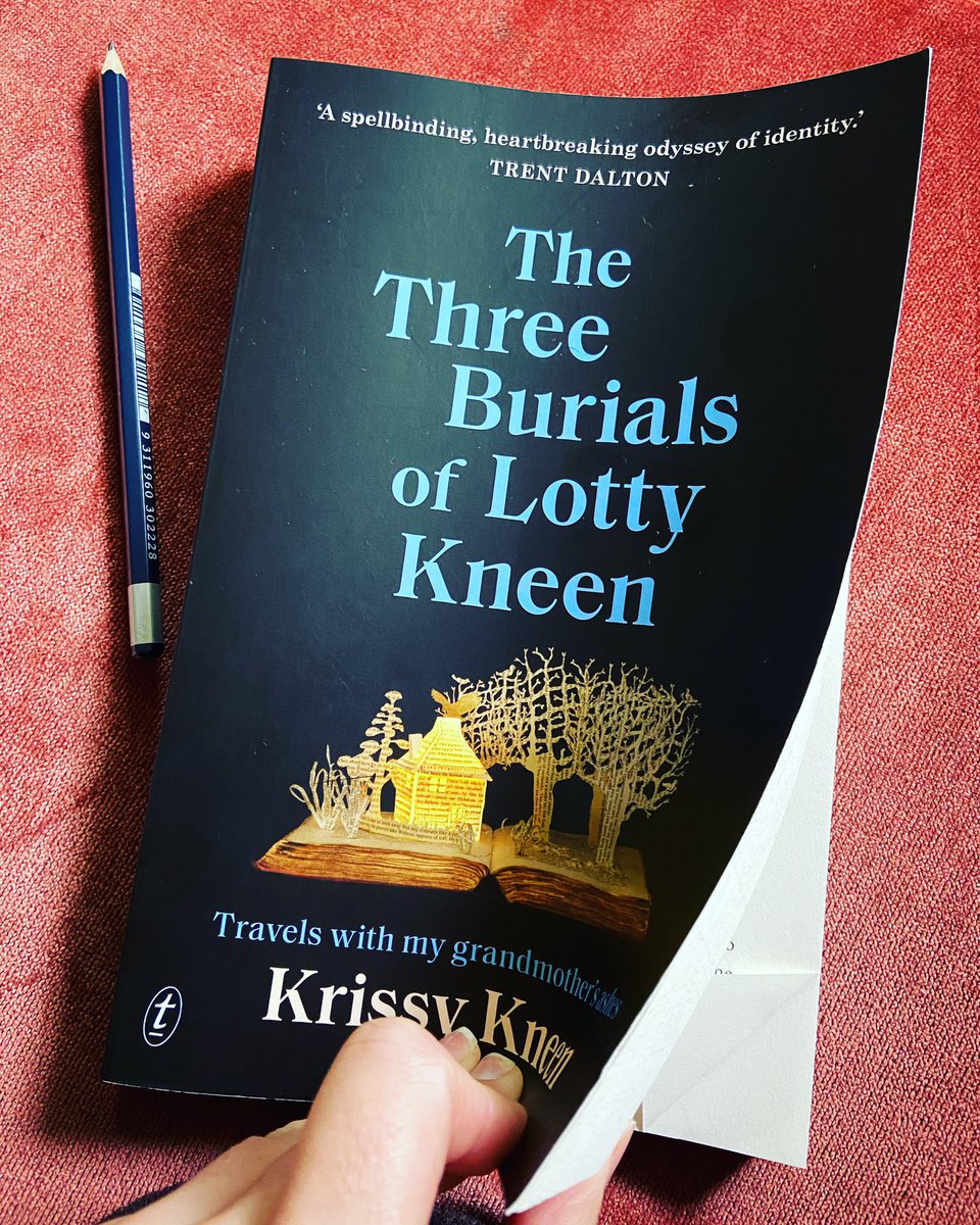 My hand opens The Three Burials of Lotty Kneen to a dog-eared page. On the cover is a paper house emerging from an open book. The book sits on my rose pink couch with a pencil beside it.