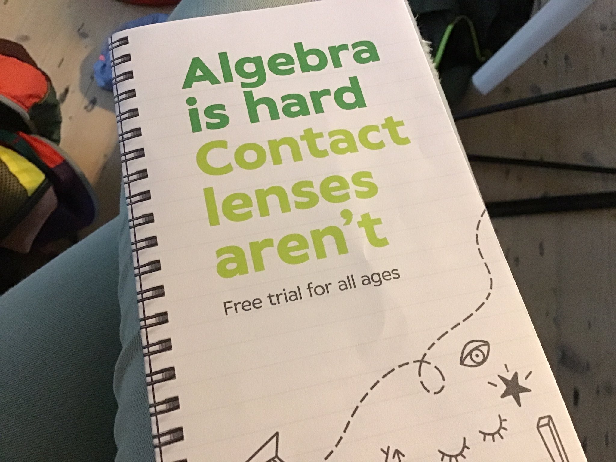 Photograph showing a pamphlet in Specsavers branding colours with the text "Algebra is hard / Contact lenses aren't // Free trial for all ages" resting on someone's knee