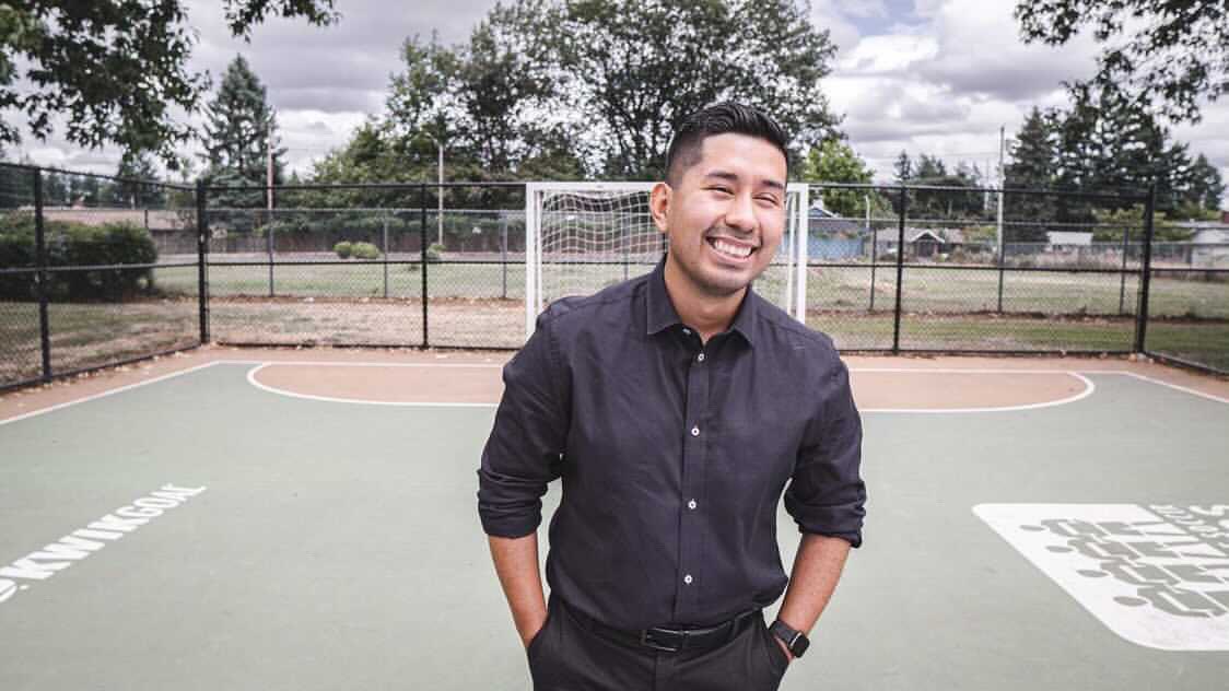 Ricki standing in the middle of an outdoor futsal court. He is dressed in black pants and a black button up shirt with the top button undone and the sleeves rolled up. He has his hands in his pockets and has a charming smile in his face.