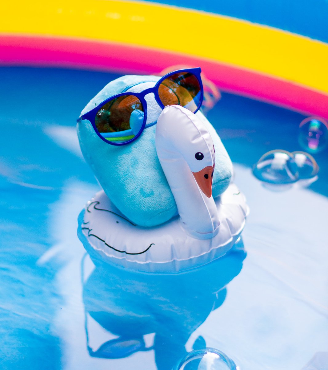 A cyan Crewmate plushie on a small swan floaty. It's wearing sunglasses, looking very chill. Both are in a kiddie pool filled with water.