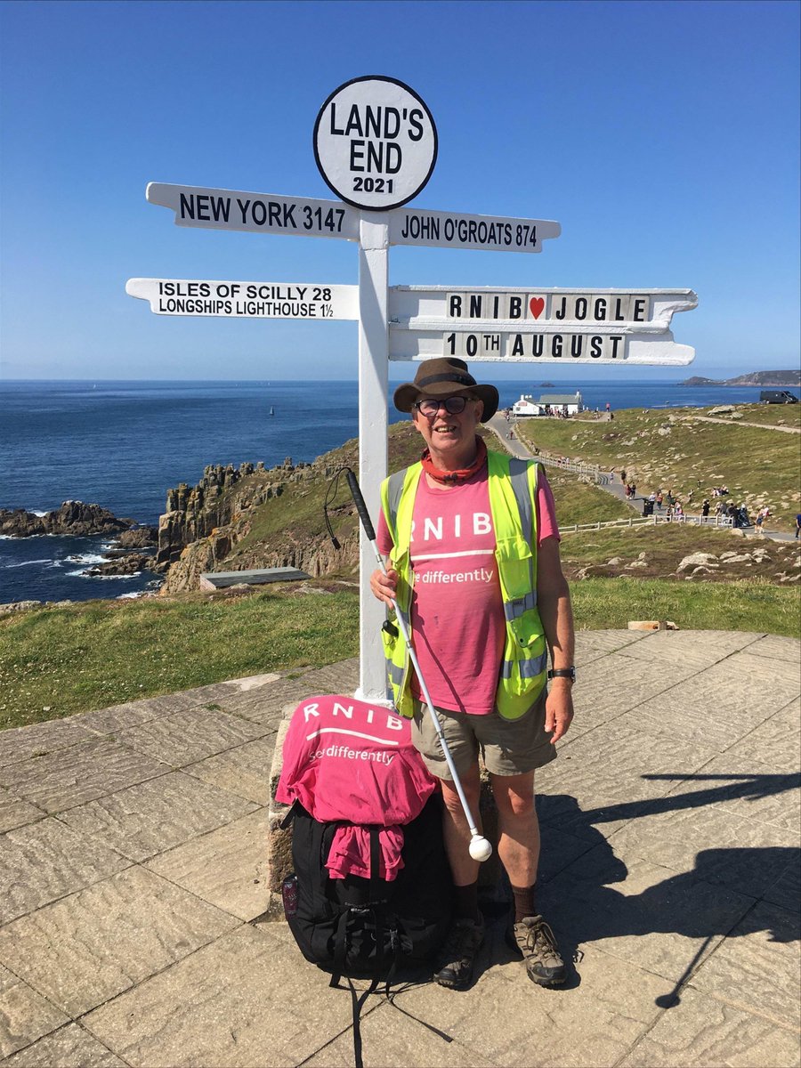 Richard standing in front of the Land's End sign. He is holding his cane and is wearing a hat, an RNIB t-shirt and his RNIB bag is on the ground. The sign says LAND'S END 2021 at the top. Underneath it has directions to NEW YORK 3147, JOHN O'GROATS 874 and ISLES OF SCILLY 28 LONGSHIPS LIGHTHOUSE 1 1/2. Pointing to the right are the directions RNIB [LOVE HEART] JOGLE 10TH AUGUST.