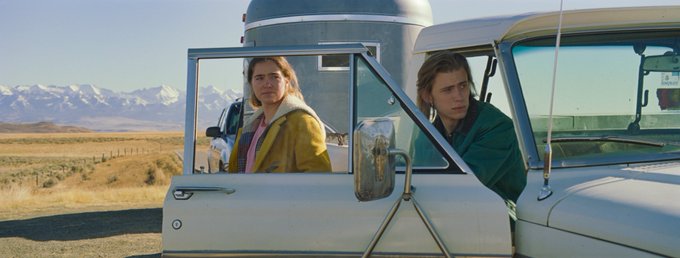Haley Lu Richardson stands next to Owen Teague sitting in a white pick-up truck with the door open, in a field with a blue silo behind them.