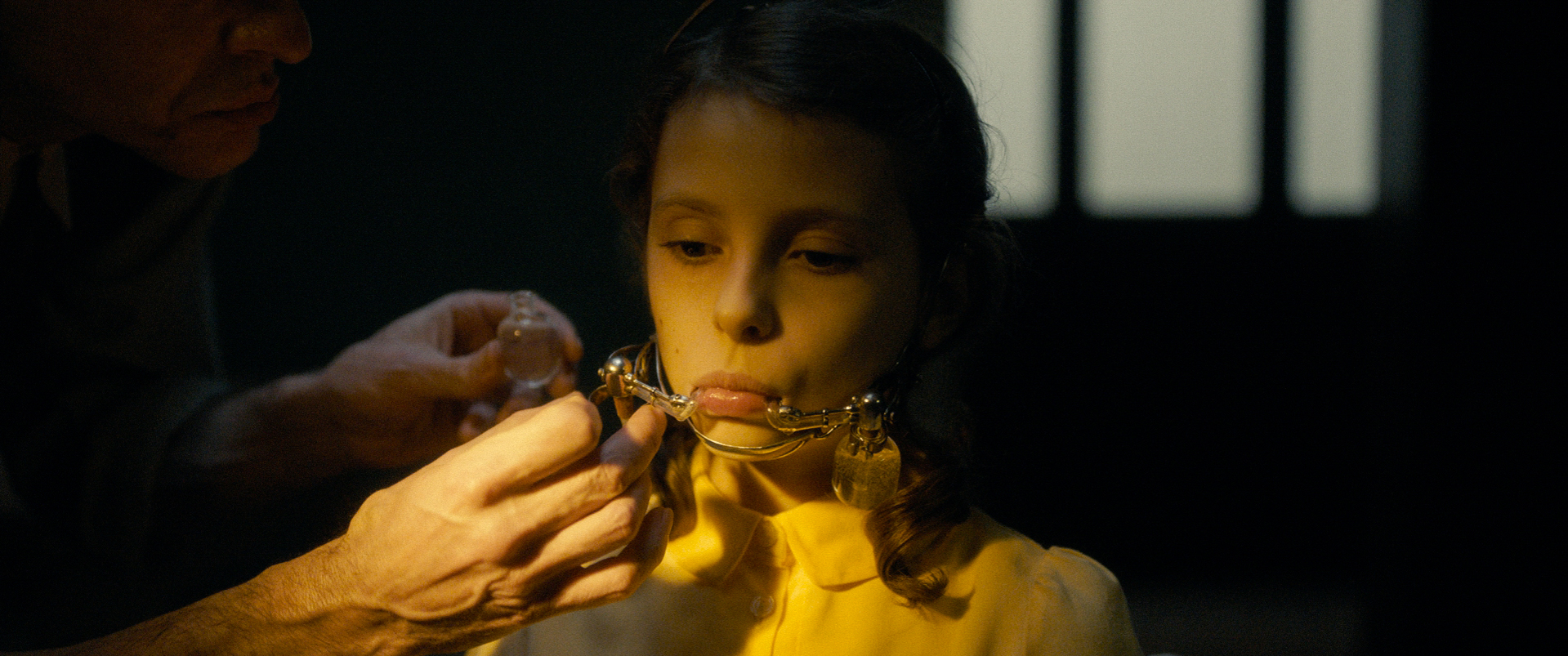 A little girl in a white dress sits in a dim room as an older man fixes a dental contraption onto her face.