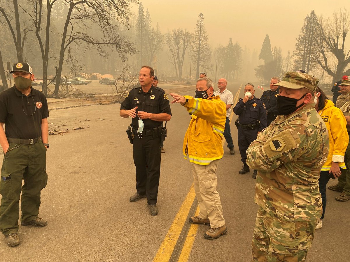 Fire service, national guard, police, forest service standing on road in smoke in burnt out landscape talking.