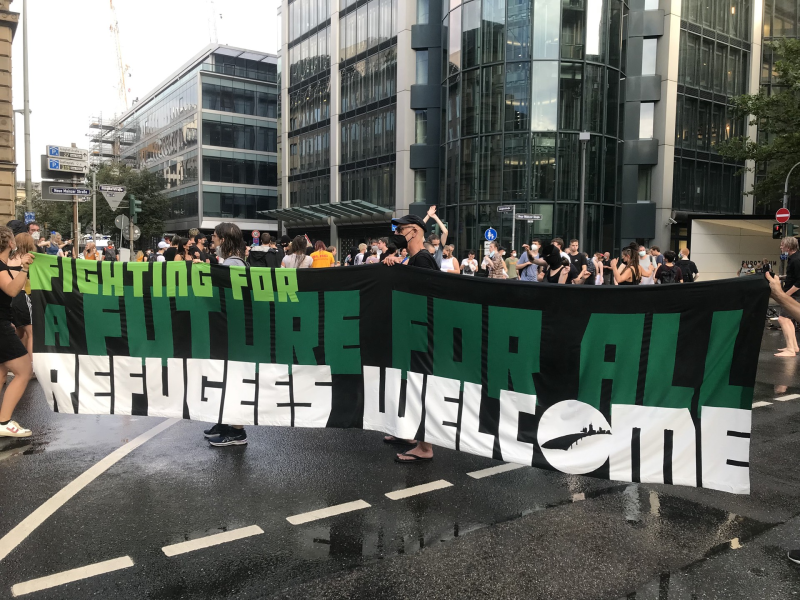 FIGHTING FOR<br>A FUTURE FOR ALL<br>REFUGEES WELCOME<br>Seebrücke-Transpi mit Aktivisti an der NeueMainze/Junghof, dahinter tanzende Menschen.