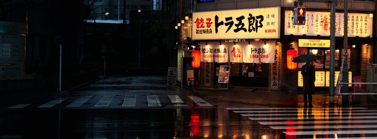 Tokyo Japan. A man waits at a traffic light holding an umbrella. Behind him is a gyoza restaurant with lanterns reflecting in the rain. The left side of the image is dark from no light.