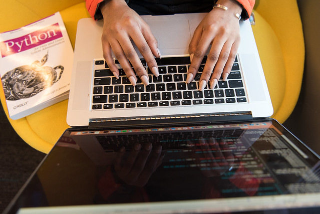 Black woman's hands on a laptop, with a Python programming book at her side. Photo CC-BY 2.0 WoC in Tech Chat