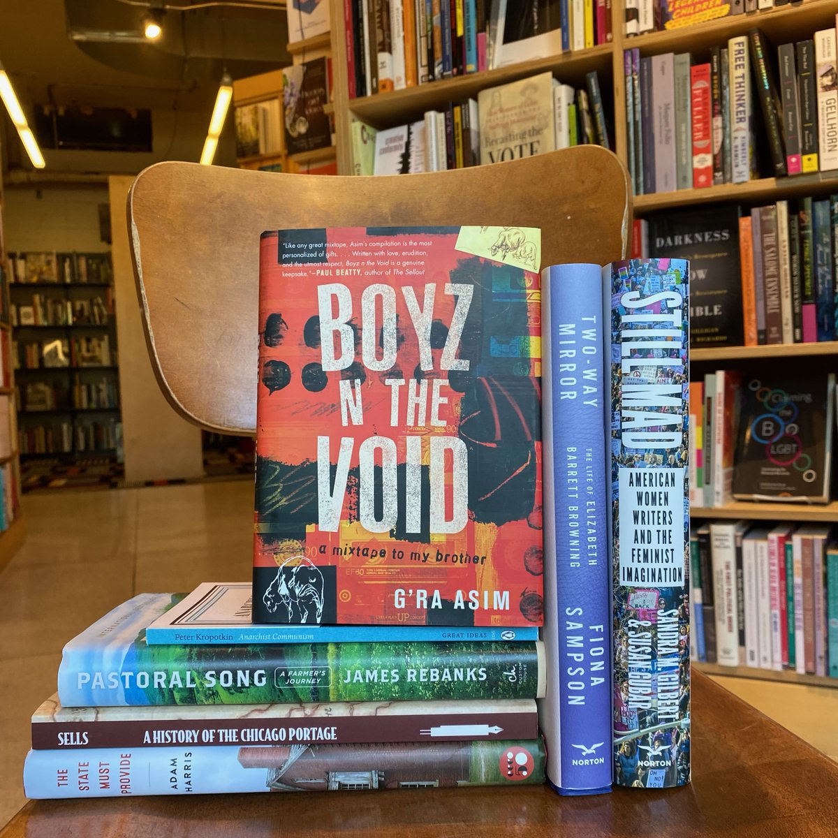 A stack of seven books in front of Seminary Co-op shelves. The books are: "Still Mad: American Women Writers And the Feminist Imagination," "Two-Way Mirror: The Life of Elizabeth Barrett Browning," "Boyz in the Void," "Anarchist Communism: Everywhere You will Find That the Wealth of the Wealth Springs From the Poverty of the Poor." "Pastoral Song," "A History of The Chicago Portage," & "The State Must Provide"