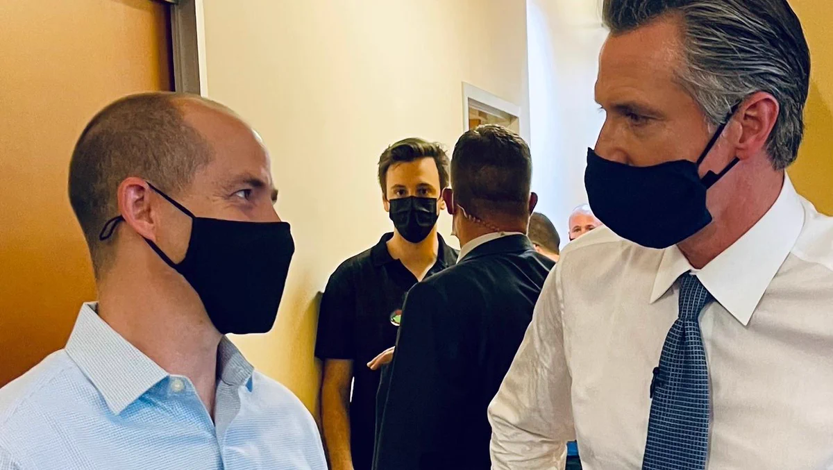 Color photograph of Rusty Hicks and Gavin Newsom looking at each other and wearing black face masks while in a hallway.