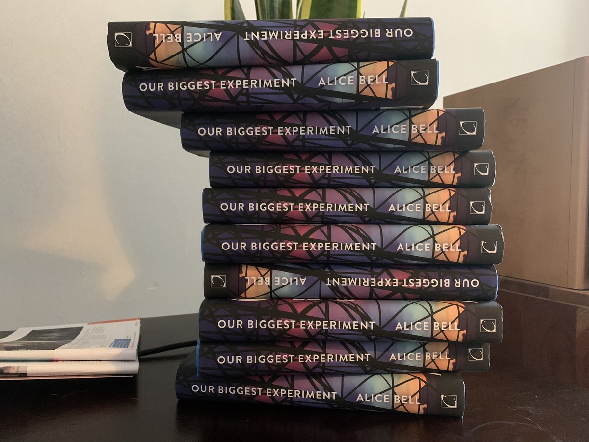 A pile of 12 hard back books on a desk: Our Biggest Experiment, Alice Bell