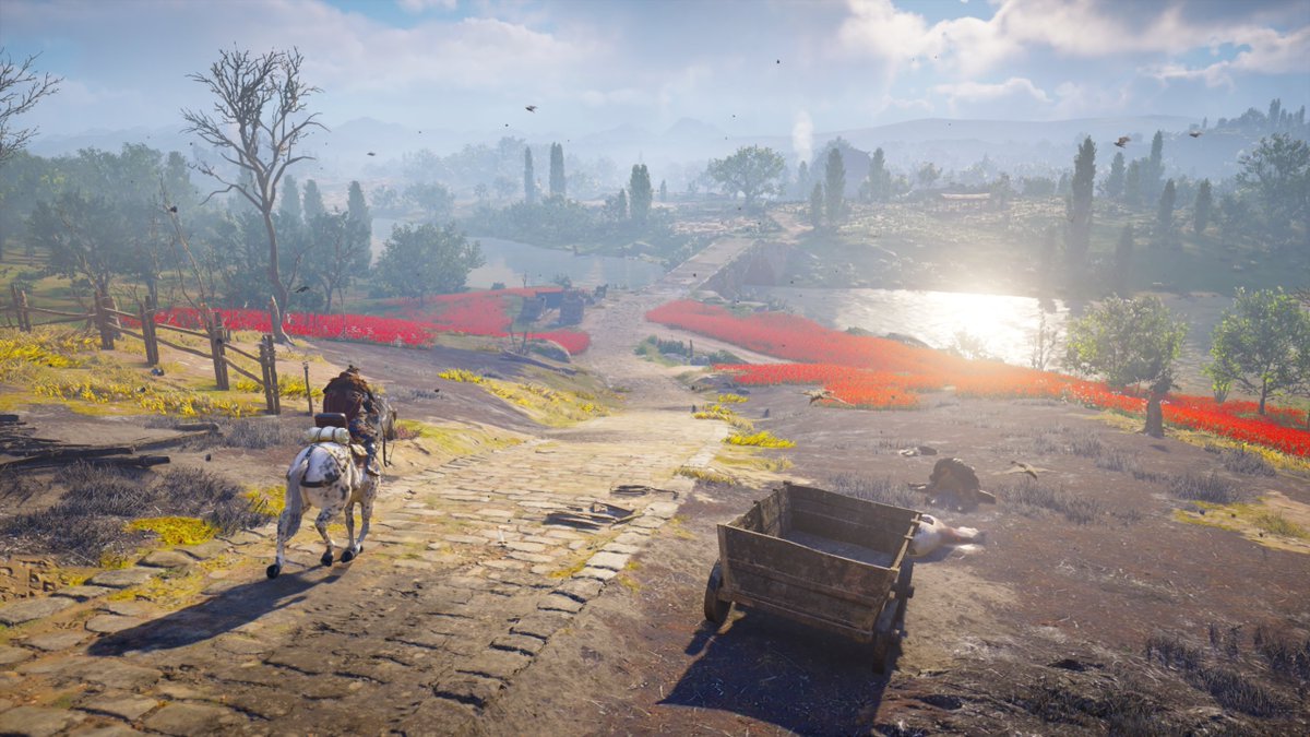 Eivor rides a horse on a cobbled pathway towardsa a field of red flowers and over a bridge, the sunlight glinting off of the water. 