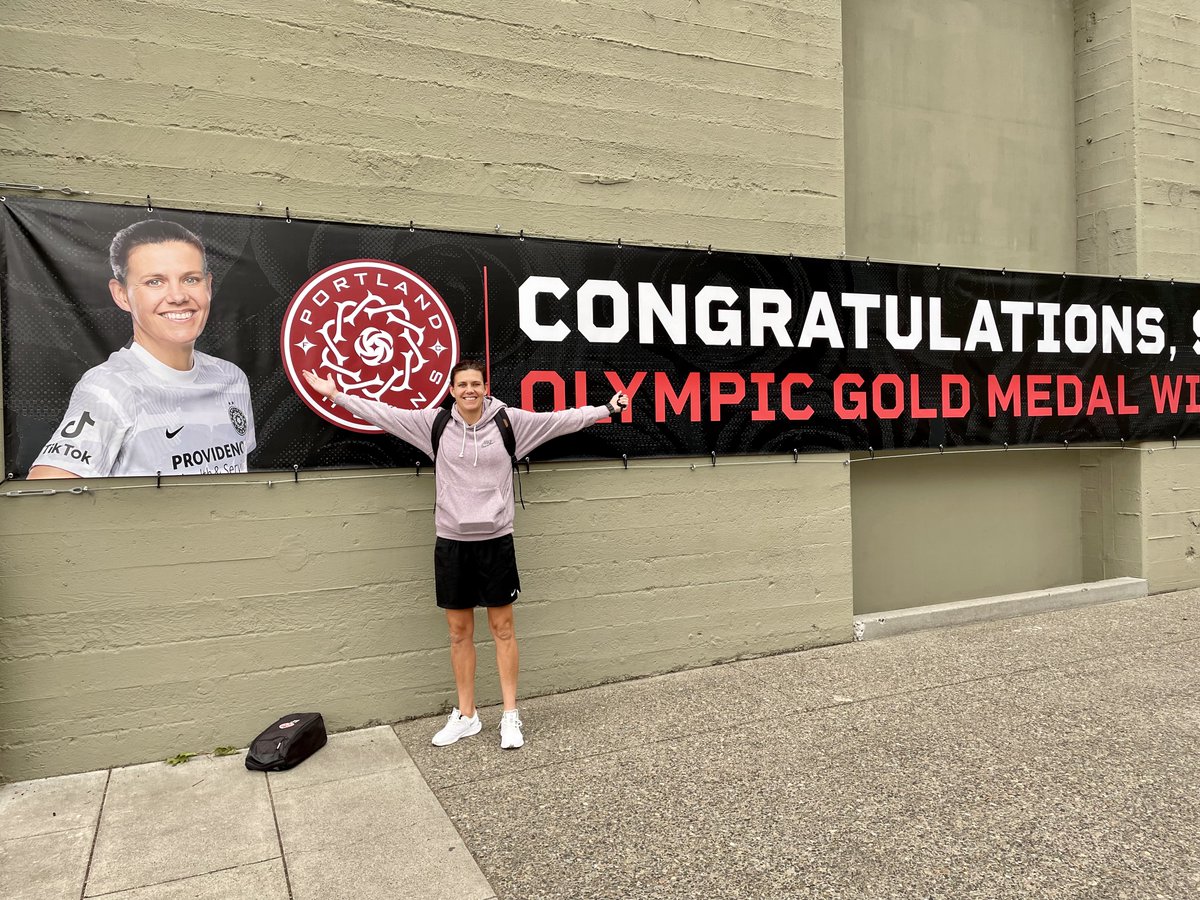 Christine Sinclair standing in front of a banner smiling. The banner has her face on it and reads "Congratulations, Sinc. Olympic Gold Medal Winner"