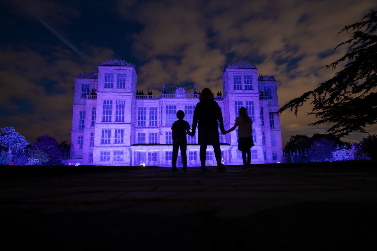 Silhouette of an adult and two children standing in front of the Elizabethan house at Hardwick which has been illuminated with purple light
