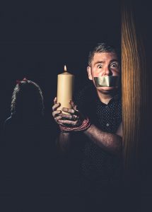 image of Darren holding a large candle with his wrists bound and sliver tape over his mouth.