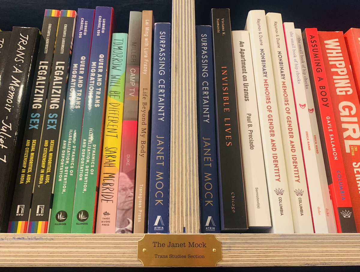 Shot from below, a photo of the “The Janet Mock Trans Studies Section” plaque with Janet Mock’s book Surpassing Certainty directly above, joined by Queer and Trans Migration, Tomorrow Will Be Different, An Apartment On Uranus, Whipping Girl, etc