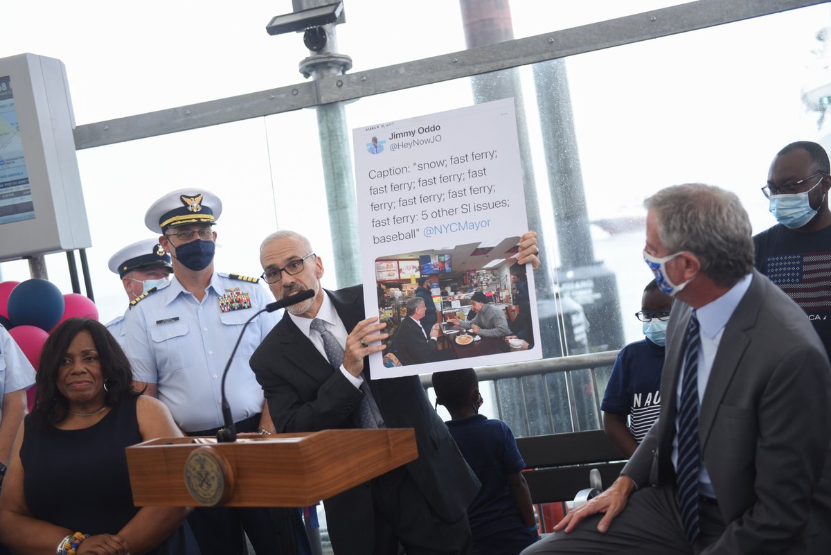 A photo of Staten Island Borough President Jimmy Oddo standing beside the Mayor at a podium and holding up a poster of a tweet showing a photo of the two of them having a conversation and the text reads "snow, fast ferry, fast ferry, fast ferry, fast ferry, fast ferry, 5 other SI issues, baseball. @NYCMayor.