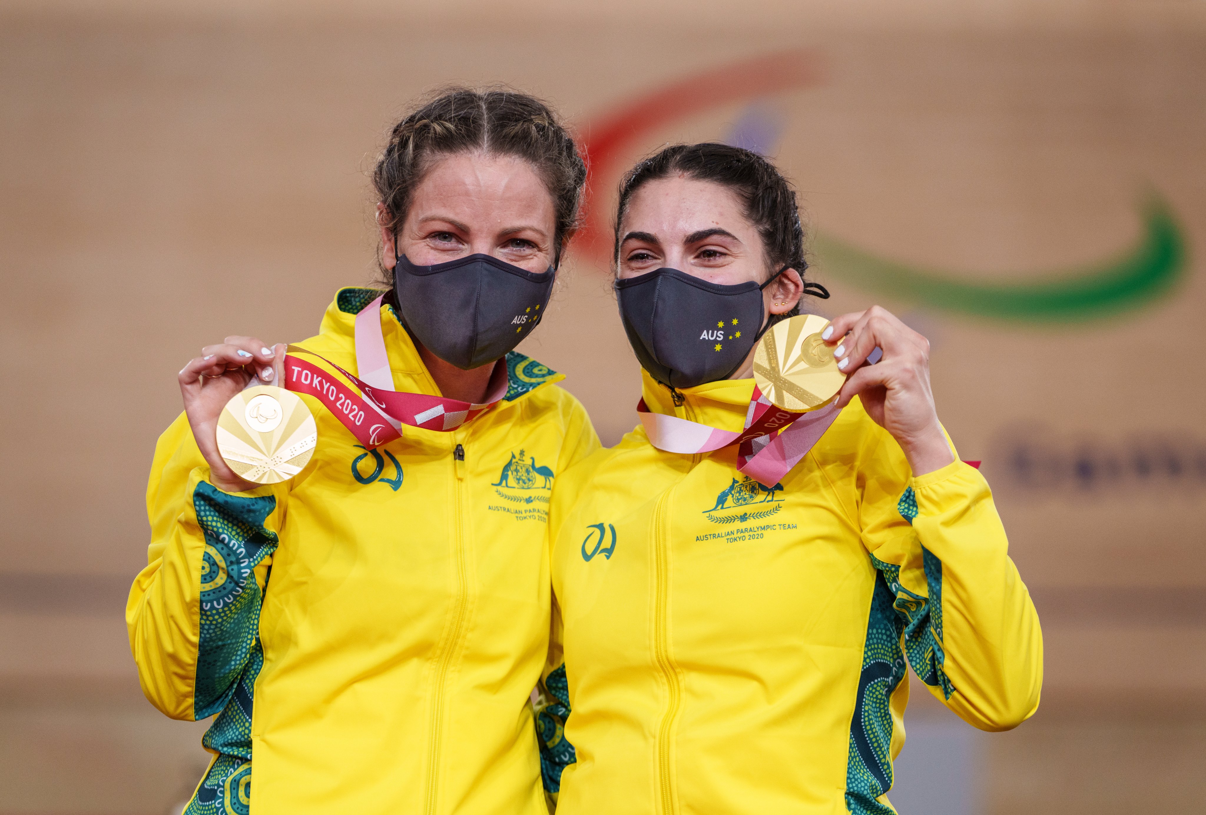 Emily Petricola and Paige Greco stand together on the podium, holding up their gold medals and smiling beneath masks. 