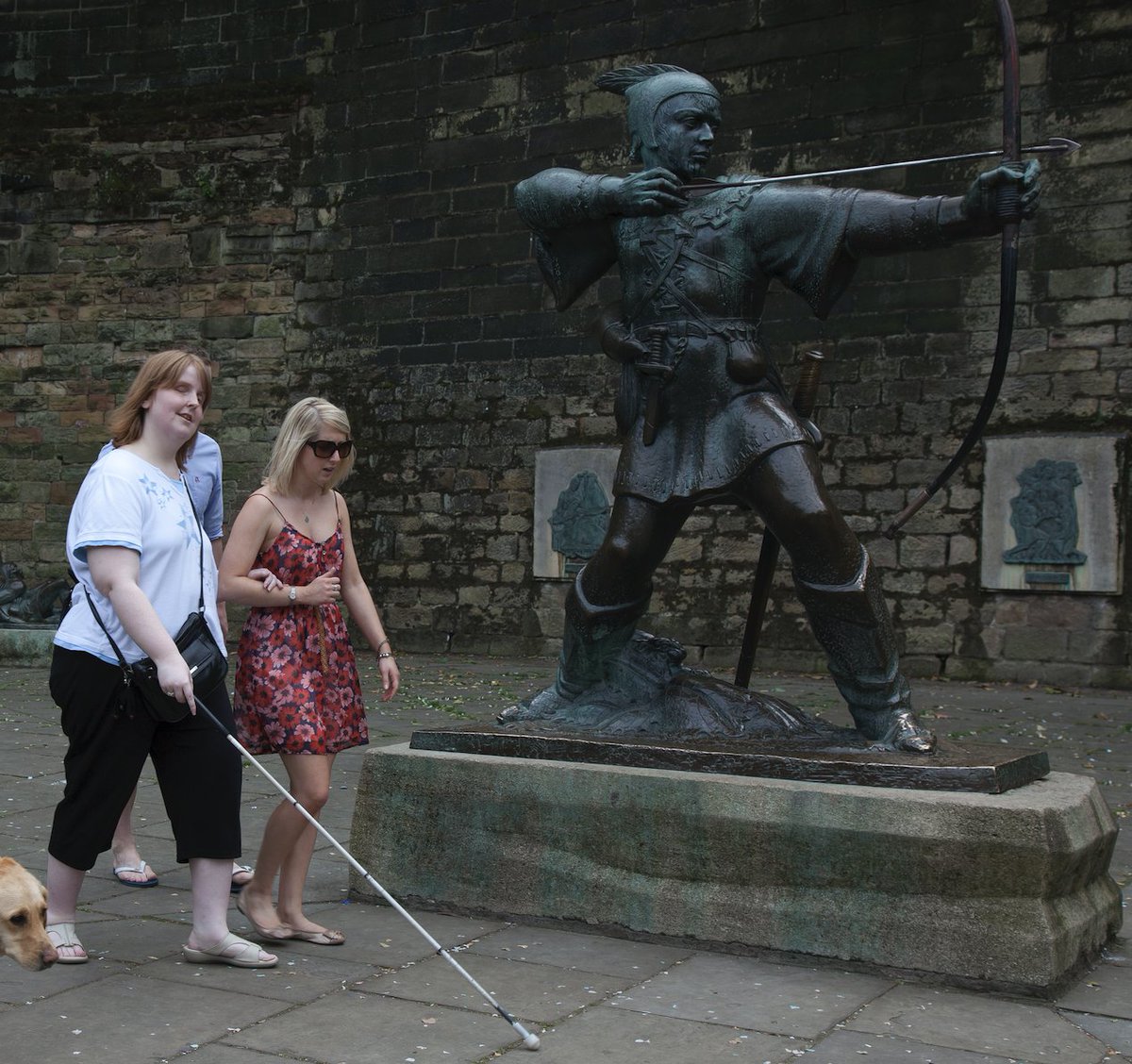 Photo shows a visually impaired woman with a white cane, being guided by one of our amazing #Volunteers past the statue of #RobinHood in Nottingham city centre.