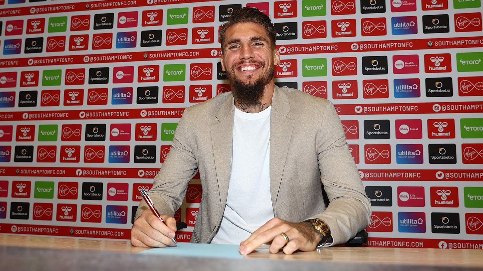 Lyanco, wearing a light-coloured blazer and white T-shirt, smiles as he signs his contract in front of the sponsors' backdrop.