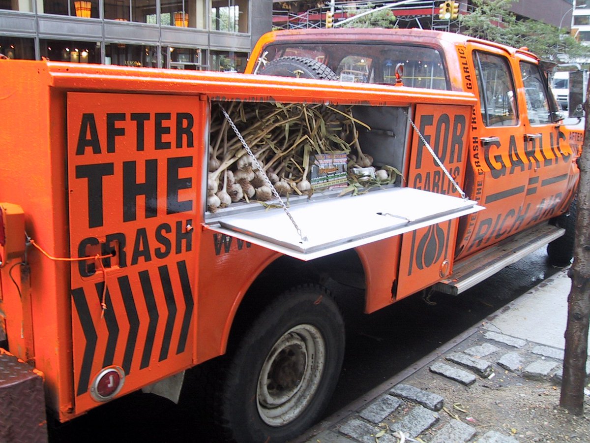 A truck loaded with cloves of organic garlic, adorned with the words 'AFTER THE CRASH' and 'GARLIC=RICH AIR', parked on a New York street.
