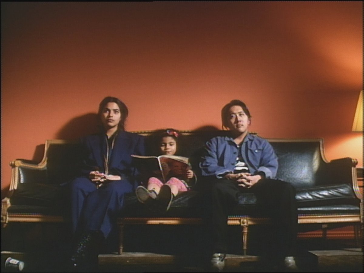 A child sits on a leather sofa between two adults in a domestic setting as they stare blankly towards the camera.