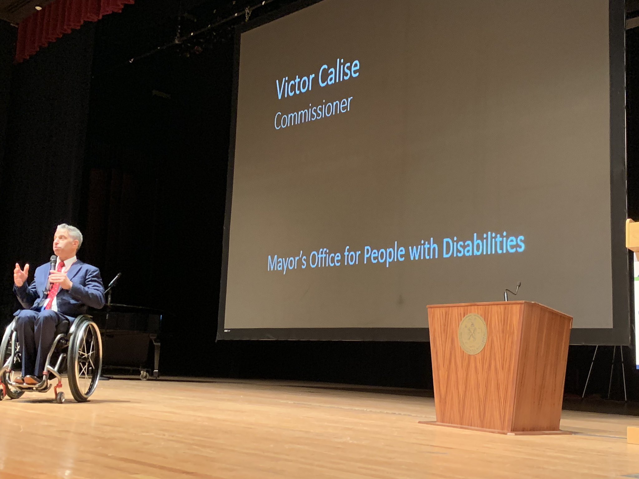 Victor Calise in a wheelchair on stage speaking beside a wheelchair podium