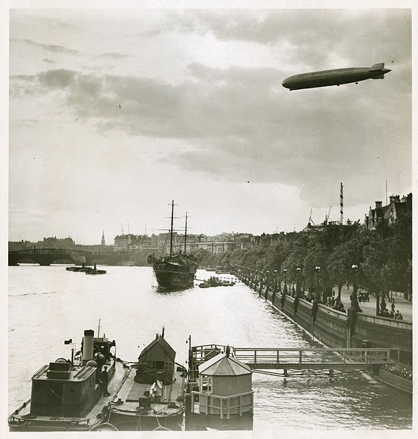 airship high in the sky over a ship in a body of water lined with trees