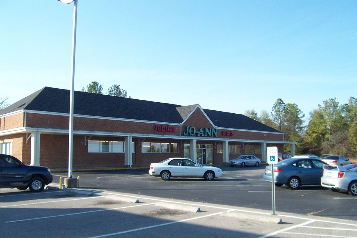 A photo of Jo Ann Fabrics shot from across the parking lot. There are a number of vehicles parked here. There is a lamp post on the right and trees in the background.