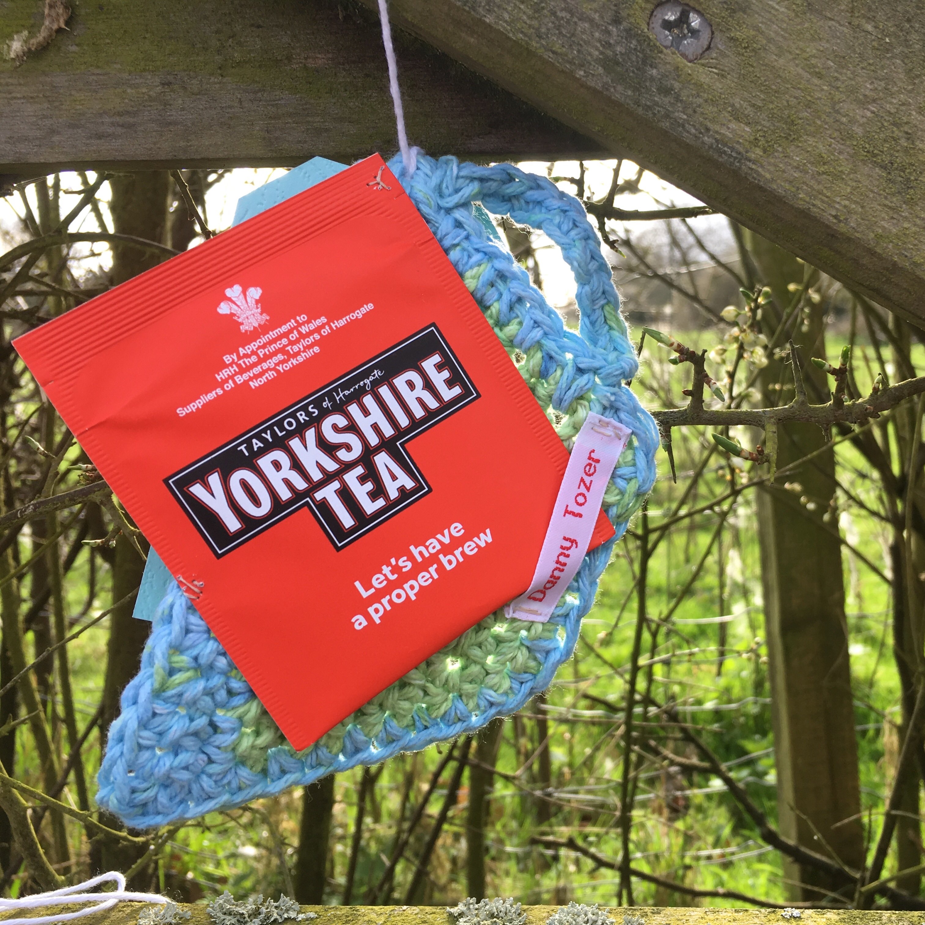 A red Yorkshire Teabag hangs on a crocheted teacup off a fence