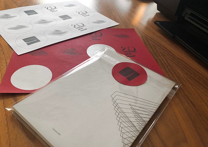 Back of an A5 zine in a clear cover, sealed with a red circular sticker with 01 on it. Other sticker sheets in the background.
