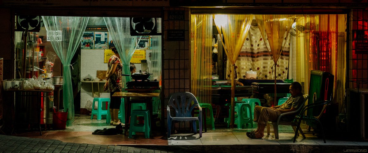 A photography series by Liam Wong - Chongqing after dark - the largest proper city in the world with a population of 30 million. In this photograph is a wide image of two shop fronts, on the left is two women and on the right is a man who seems to be sleeping in a chair.