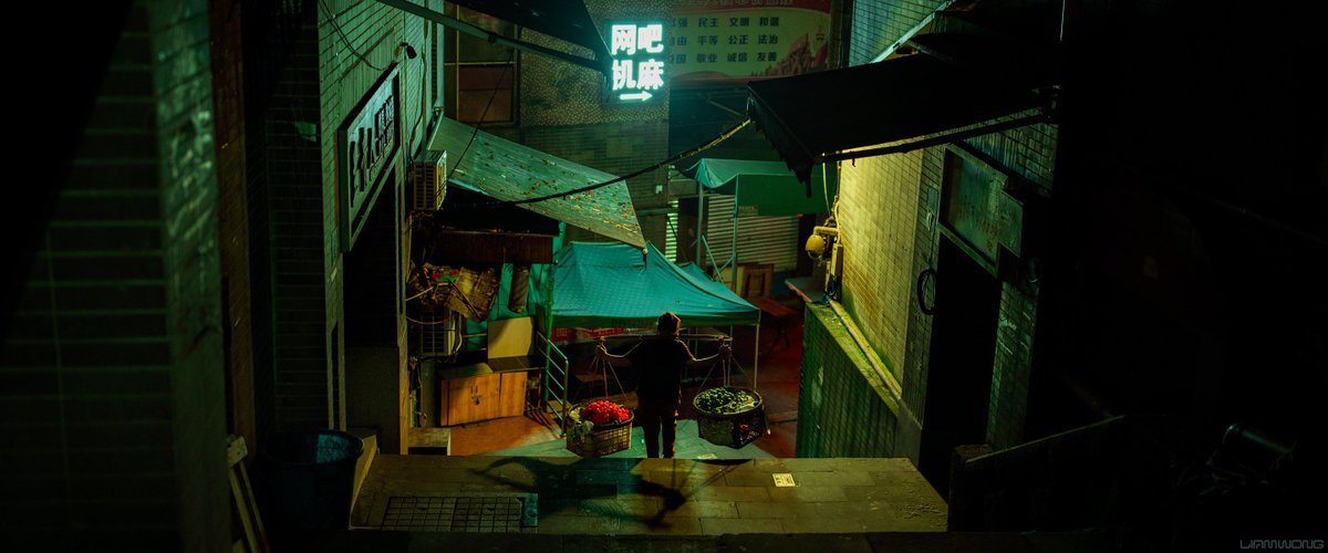 A photography series by Liam Wong - Chongqing after dark - the largest proper city in the world with a population of 30 million. In this photograph an elderly woman walks down an alley holding a bamboo stick across her shoulders, with fruit balanced on either side, later to be sold. 