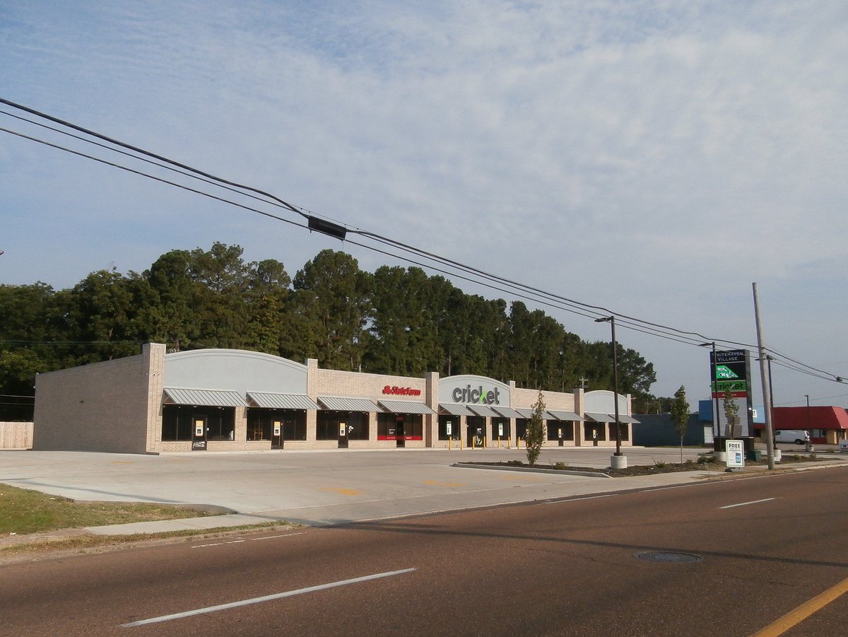 A photo of a strip mall with a Cricket Wireless store on a four-lane road. There are no cars in the parking lot. Powerlines cross the frame and there are trees and other commercial buildings in the background.