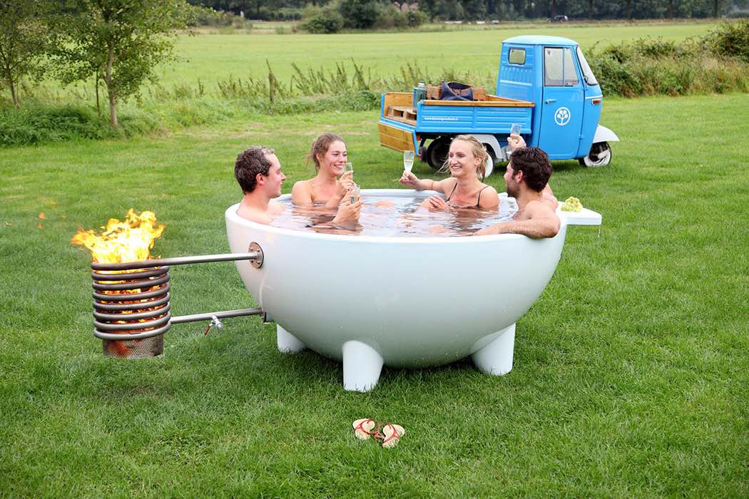 Four naked people drinking champagne in a small portable hot-water tub with a new wood-burning attachment on a lawn.