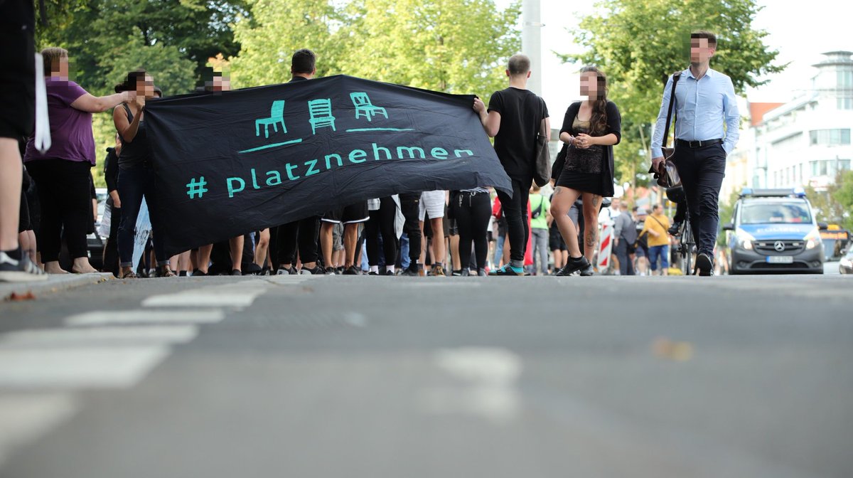 Junge Leute halten ein Transparent des Bündnis Platznehmen, auf das Stühle gemalt sind. Im Hintergrund steht ein Polizeiwagen.