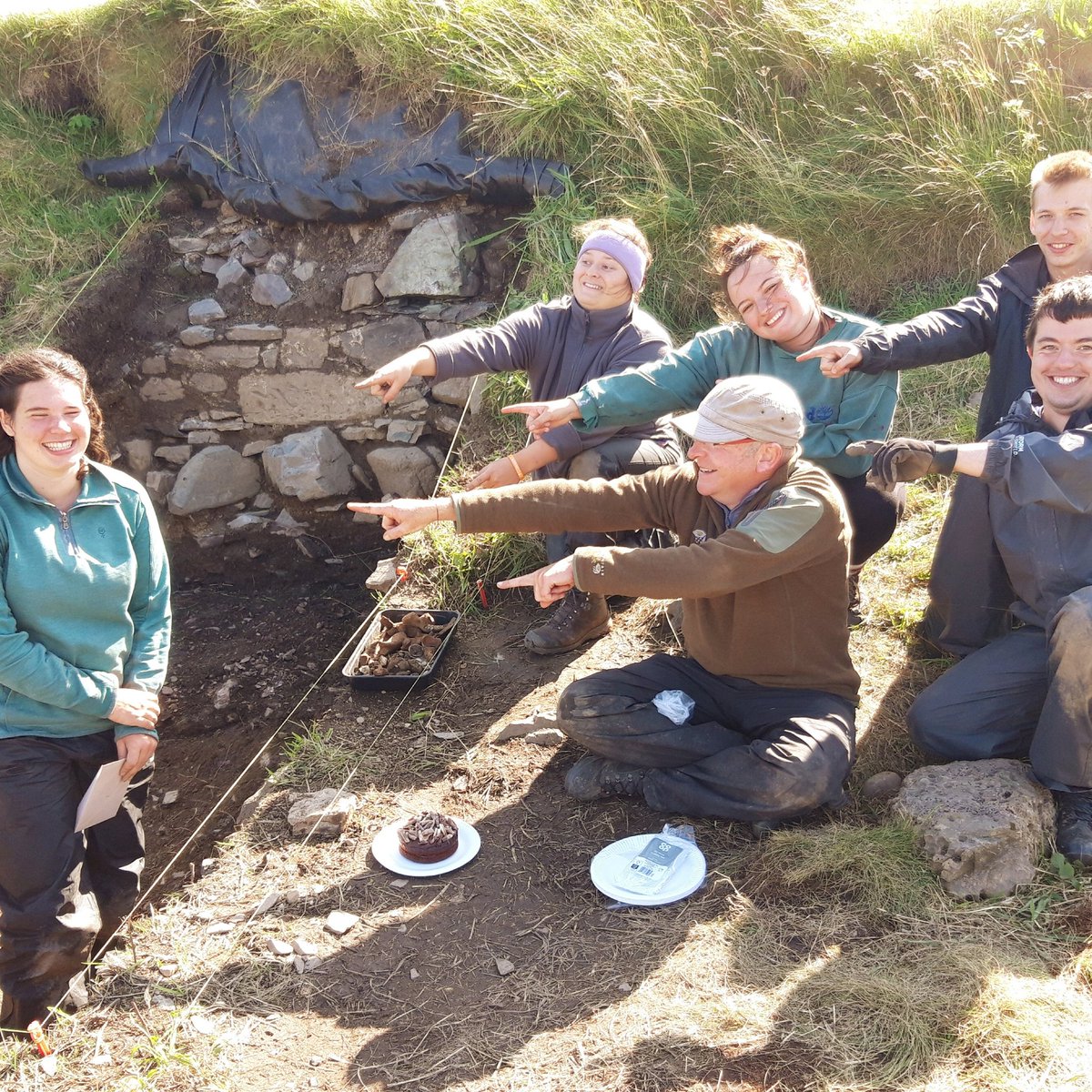 islayheritage's tweet photo. It's Victoria's 20th birthday in Trench 1b #dunyvaigdig. Meanwhile in the trench the deposit looks a bit like nutty chocolate cake mix but, but they have excavated the last of the midden containing lots of butchered bone, and are down to a levelling deposit pre-dating the wall. https://t.co/sfkA5cMMfy