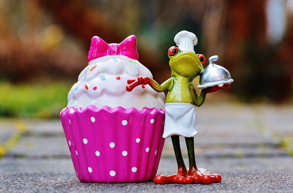 a photograph of a frog figurien wearching a chef's hat, apron, holding a tray and standing next to a cupcake figurine