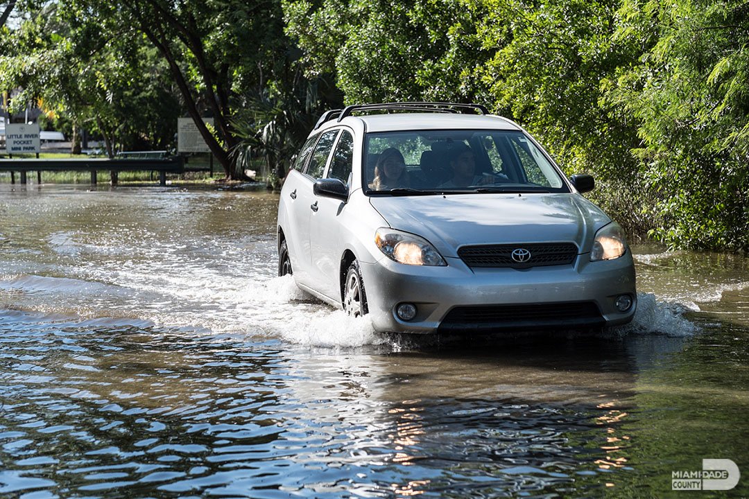 A small silver car going through a flooded street.