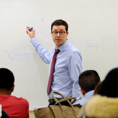 A professor wearing a tie teaches at the white board.