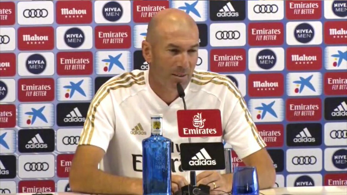 A bald man wearing a white Real Madrid Adidas shirt sits at a press conference table speaking into a microphone, with a backdrop and table displaying "Emirates," "Adidas," "NIVEA MEN," "Mahou," and Audi logos. The Spanish caption jokingly frames him as a figure ("Domenicalli") being questioned about the danger of operating in a conflict zone, humorously comparing a tense press conference setting to a war zone.