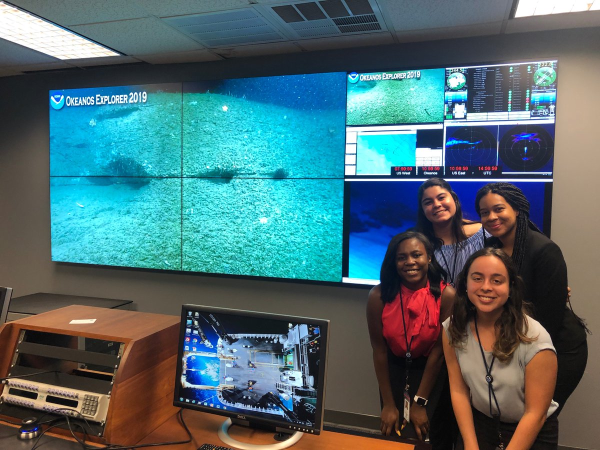 Four female students stand next to a big screen displaying ocean exploration video footage.